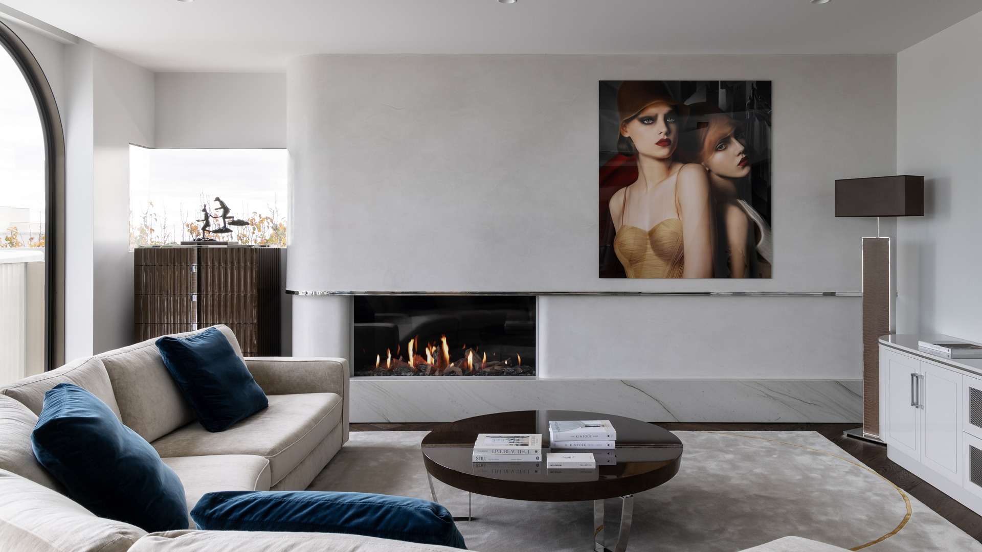Modern gas fireplace set in a curved, plaster-finished surround with marble base and chrome detailing, framed by dark herringbone timber flooring and soft neutral-toned furniture.