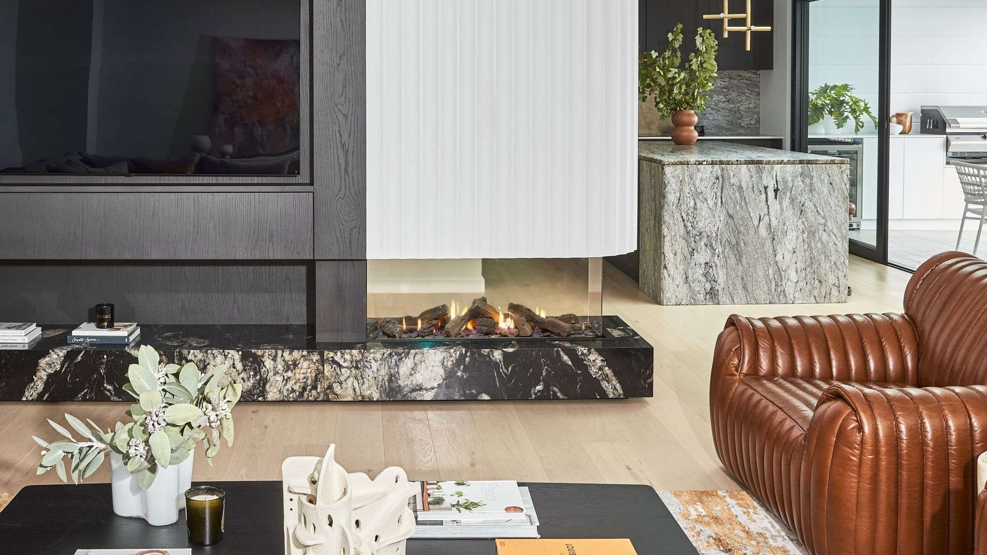 Contemporary living space featuring a glass-sided peninsula gas fireplace with dramatic marble base, fluted white cladding, rich timber cabinetry, and a bold leather armchair, connecting to a modern stone kitchen and outdoor entertaining area.